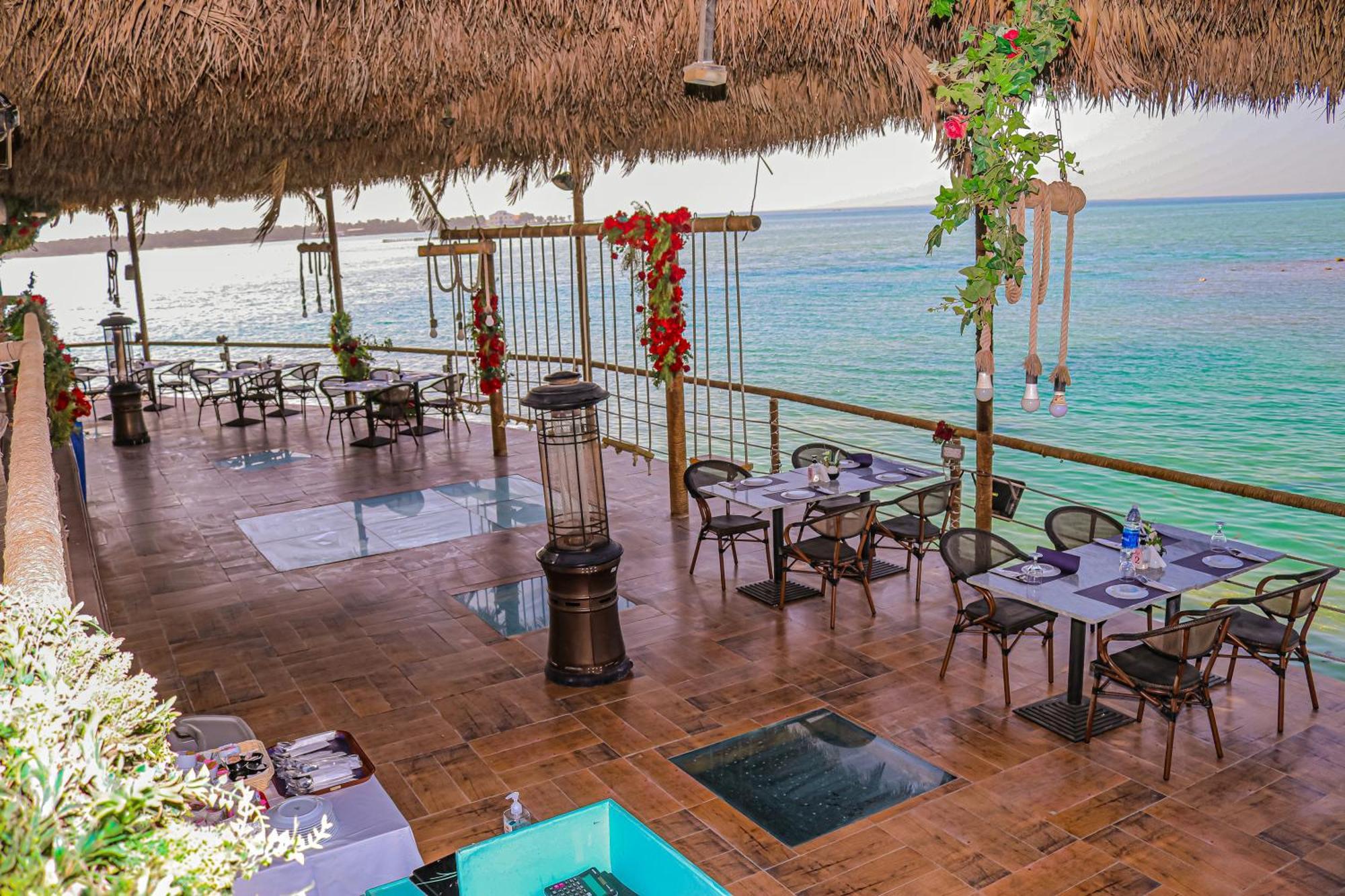 Lagoona Beach Luxury Resort And Spa Manama Eksteriør billede The photo depicts a coastal restaurant or dining area with an open-air design. It features a wooden floor and tables set for dining, surrounded by chairs. The space is partially covered by a thatched roof, providing shade while allowing for a view of