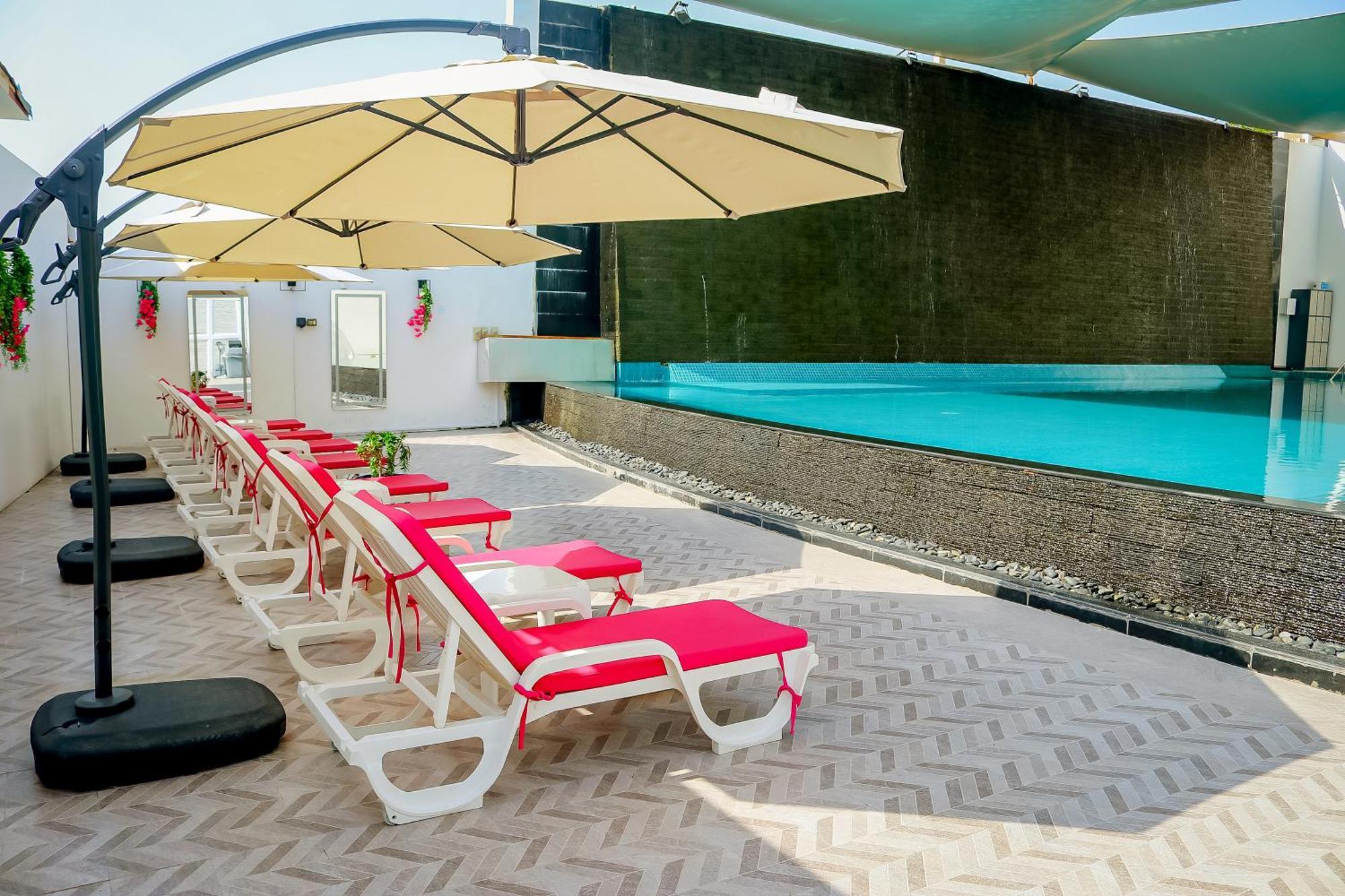 Lagoona Beach Luxury Resort And Spa Manama Eksteriør billede The photo shows a sunny poolside area with several lounge chairs arranged in a row. Each chair features red cushions and is positioned under large, beige umbrellas for shade. In the background, there is a beautiful swimming pool with clear blue water