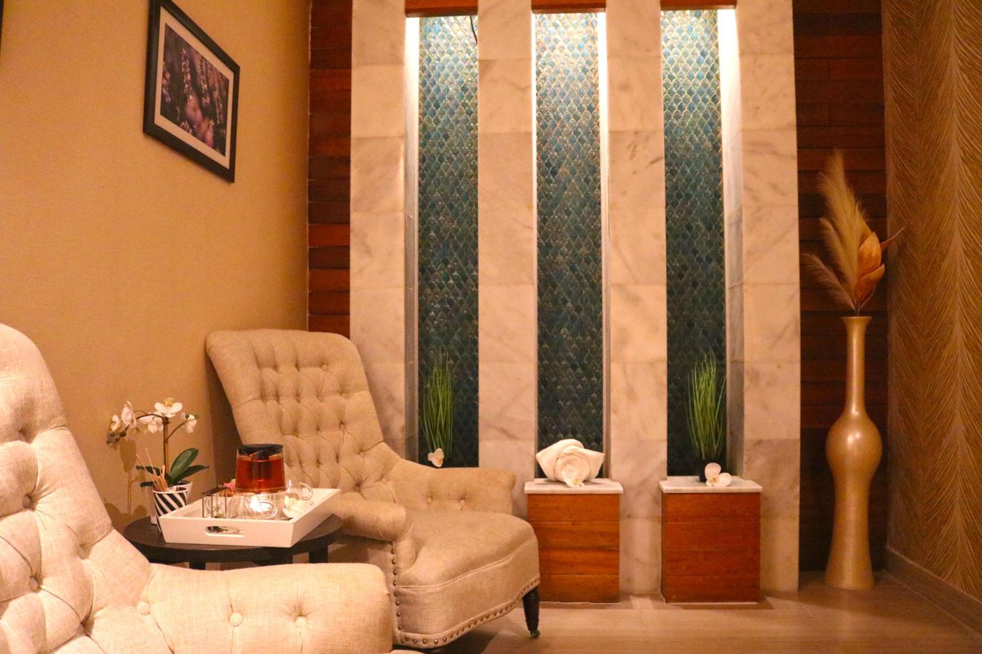 Lagoona Beach Luxury Resort And Spa Manama Eksteriør billede The image shows a relaxing interior space, likely a waiting area or lounge. There are two plush, beige armchairs positioned near a small table, which holds a glass pitcher and a cup. The background features tall, elegant windows with decorative blue 