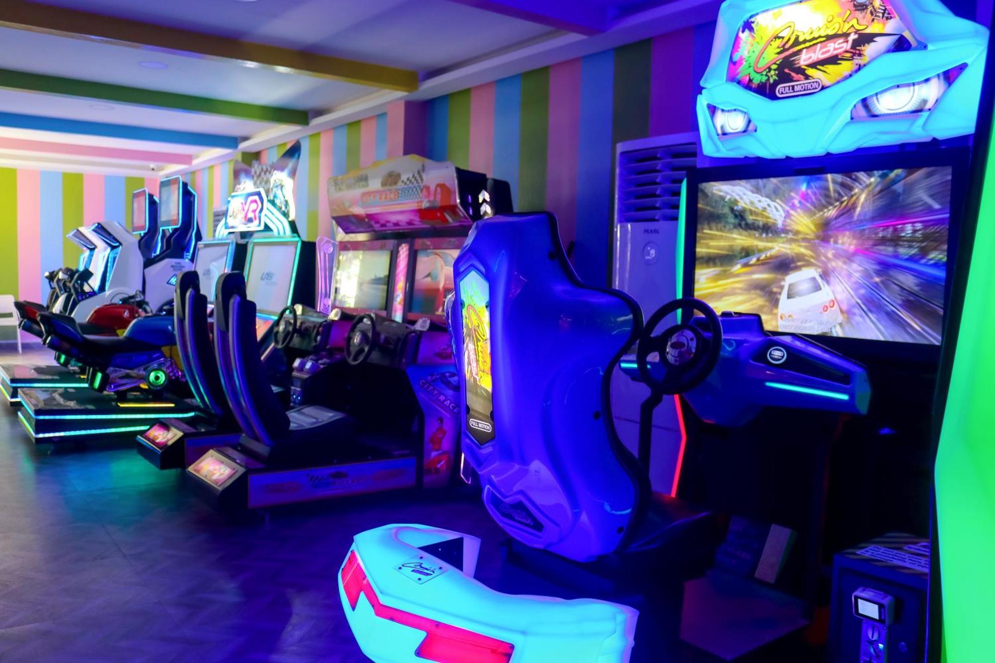 Lagoona Beach Luxury Resort And Spa Manama Eksteriør billede The photo shows an arcade area filled with various arcade machines. The setting is illuminated with colorful neon lights, creating a vibrant atmosphere. There are several racing game machines with steering wheels and seats, each accompanied by screen