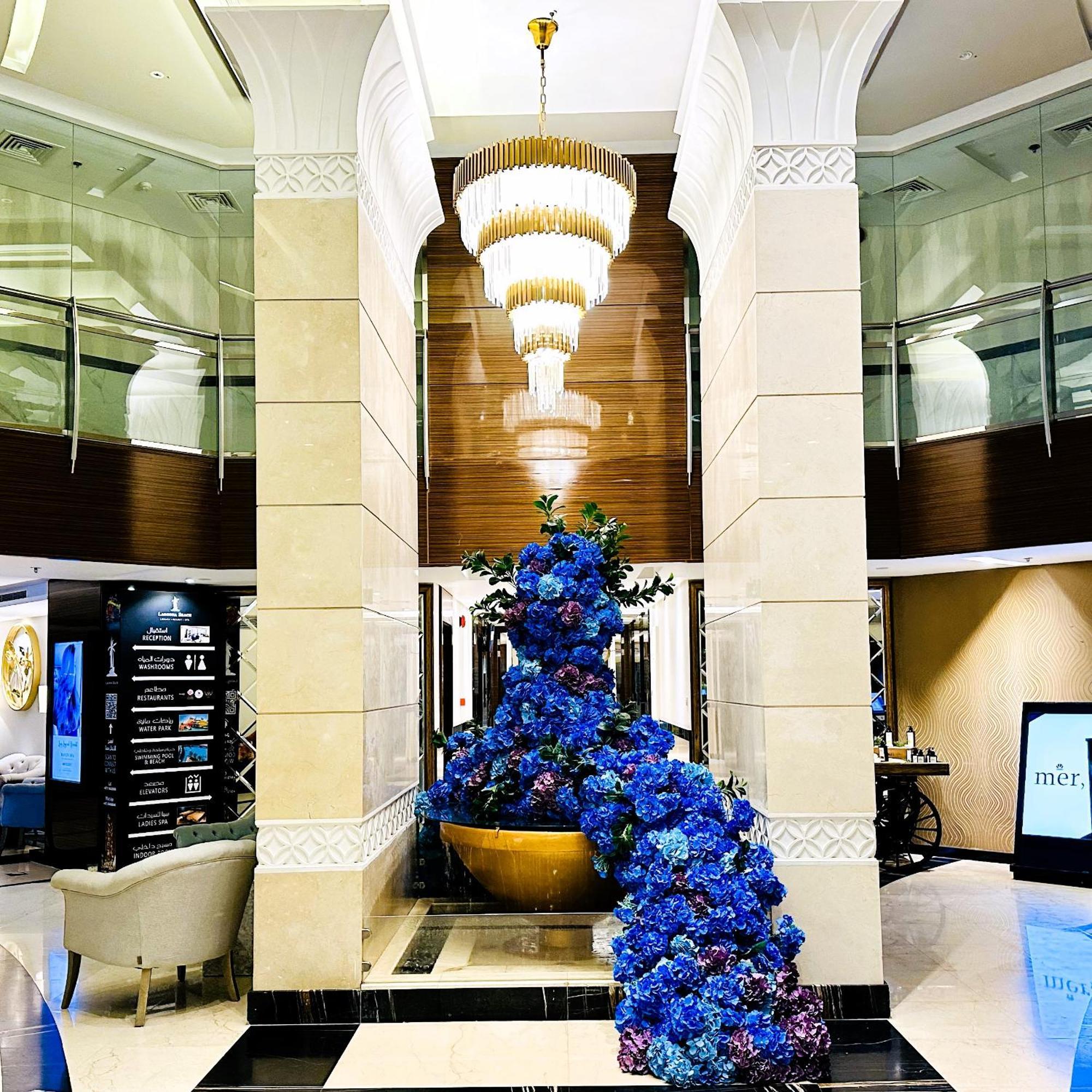 Lagoona Beach Luxury Resort And Spa Manama Eksteriør billede The photo depicts an elegant lobby or entrance area of a building. Central to the scene is a large floral arrangement featuring vibrant blue flowers. This arrangement is positioned in a decorative bowl. Above it hangs a stylish chandelier with a laye
