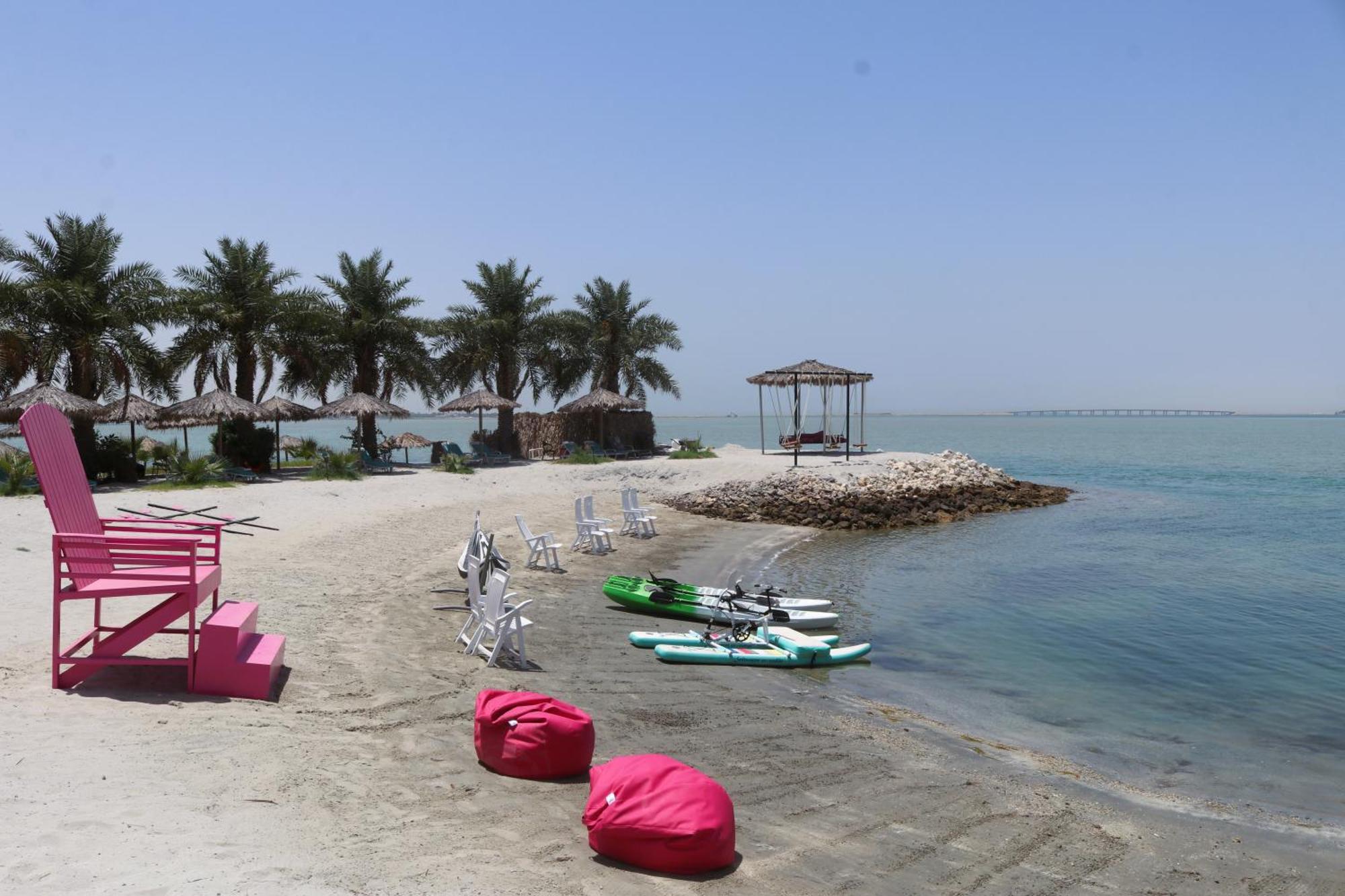 Lagoona Beach Luxury Resort And Spa Manama Eksteriør billede The photo depicts a serene beach scene with clear blue water and a sunny sky. In the foreground, there are colorful pink bean bags and a large pink chair on the sand. A few white chairs and loungers are positioned near the water's edge. To the side, 