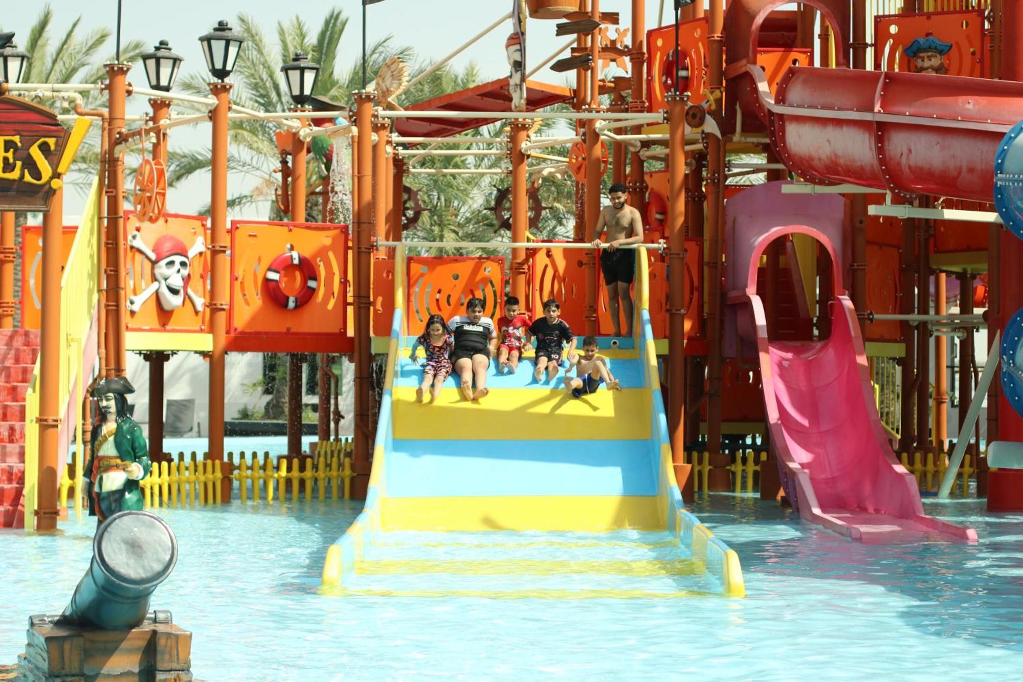 Lagoona Beach Luxury Resort And Spa Manama Eksteriør billede The photo shows a colorful water park scene featuring a play area with multiple slides. There are children enjoying themselves, sliding down bright yellow and pink slides into a pool of water. The structure has pirate-themed decorations, including a 