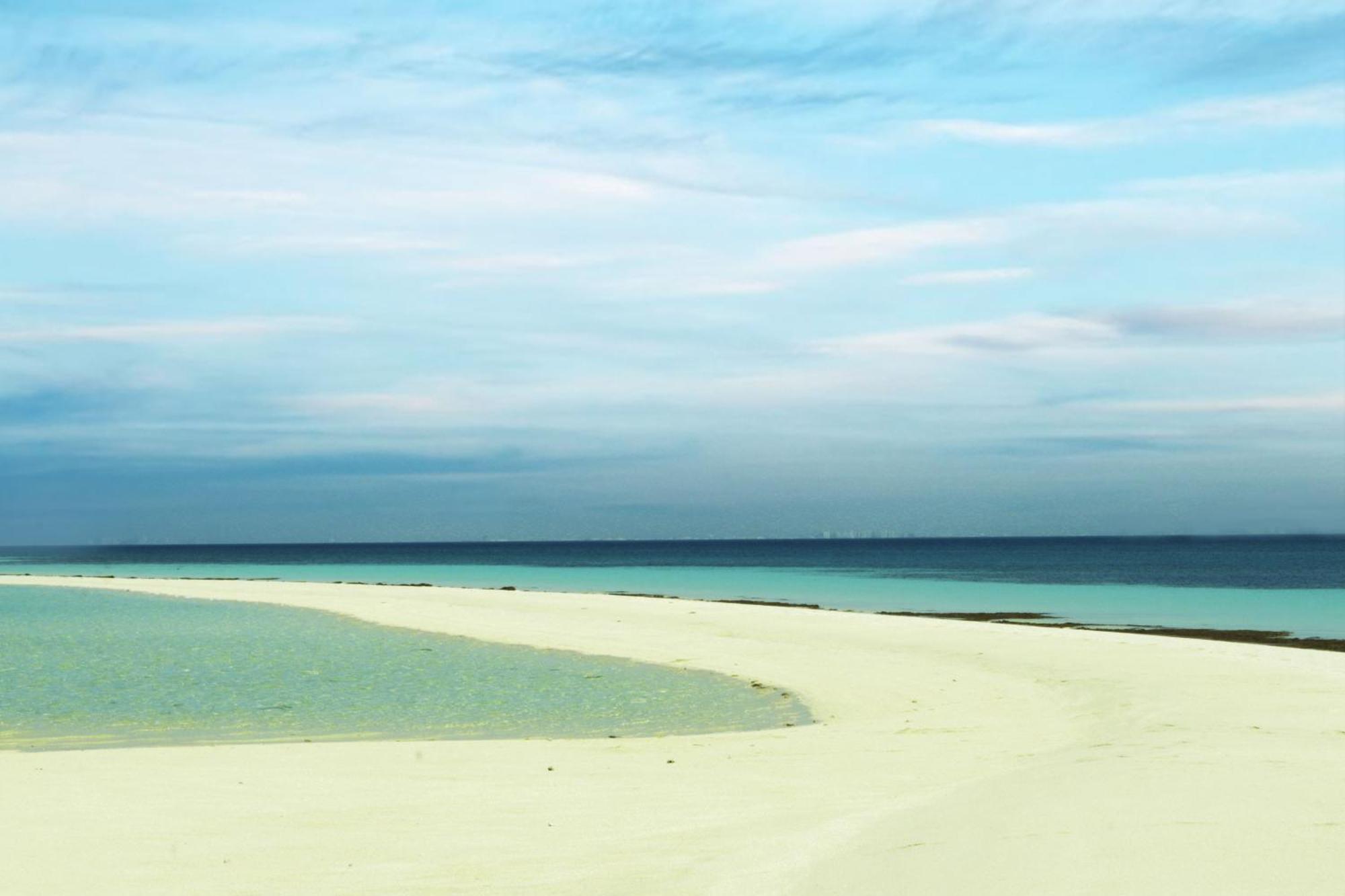 Lagoona Beach Luxury Resort And Spa Manama Eksteriør billede The photo depicts a serene beach scene with a gentle curve of sandy shore leading into shallow turquoise waters. The calm ocean stretches into the horizon, meeting a sky filled with soft clouds in various shades of blue. The overall atmosphere is tra