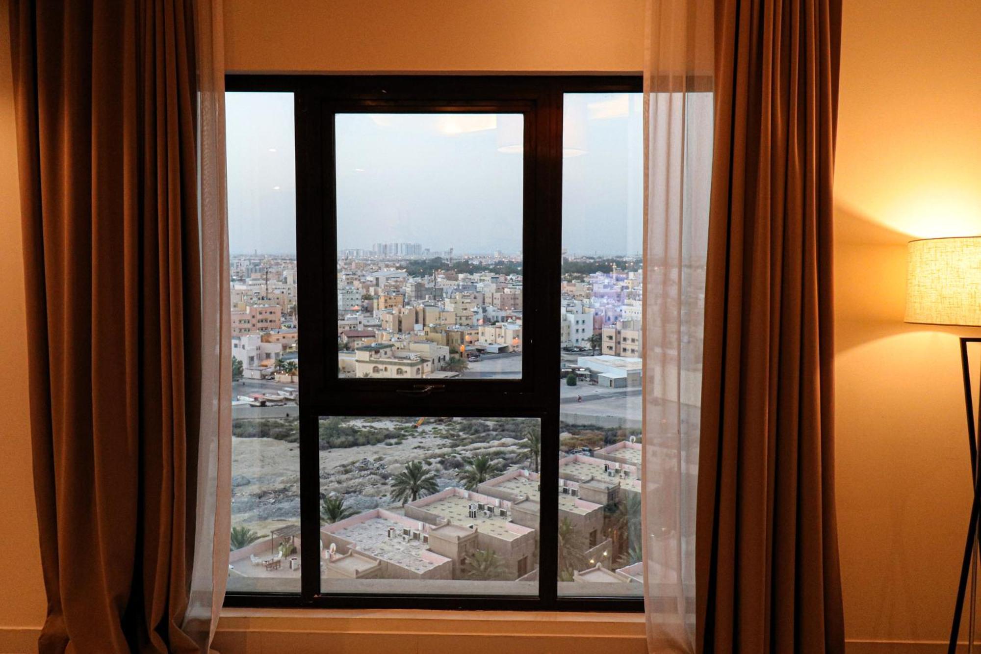 Lagoona Beach Luxury Resort And Spa Manama Eksteriør billede The photo shows a window with a black frame and sheer curtains. Through the window, there is a view of a cityscape, displaying a densely populated area with various buildings and rooftops. The sky appears slightly overcast, suggesting a muted lightin