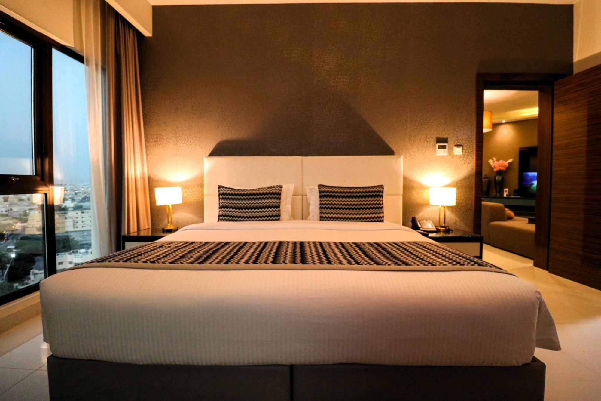 Lagoona Beach Luxury Resort And Spa Manama Eksteriør billede The photo depicts a modern hotel room featuring a large bed with a white comforter and two decorative pillows. The headboard is light-colored, and the walls are painted a dark hue. On either side of the bed, there are bedside tables with lamps. The r