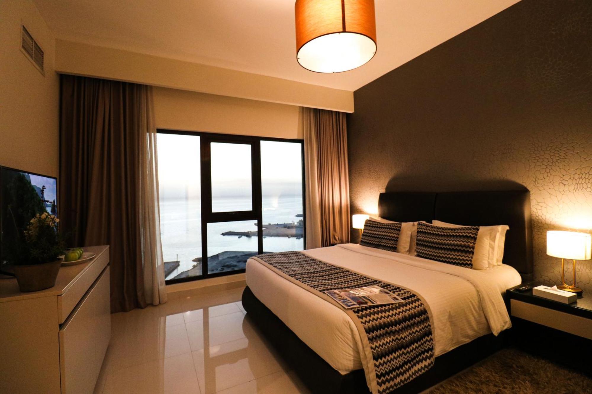 Lagoona Beach Luxury Resort And Spa Manama Eksteriør billede The photo shows a modern hotel room featuring a neatly made bed with decorative pillows. The room has a stylish design with a dark accent wall and a large window that offers a view of the outdoors. Natural light comes in, giving the space a bright an