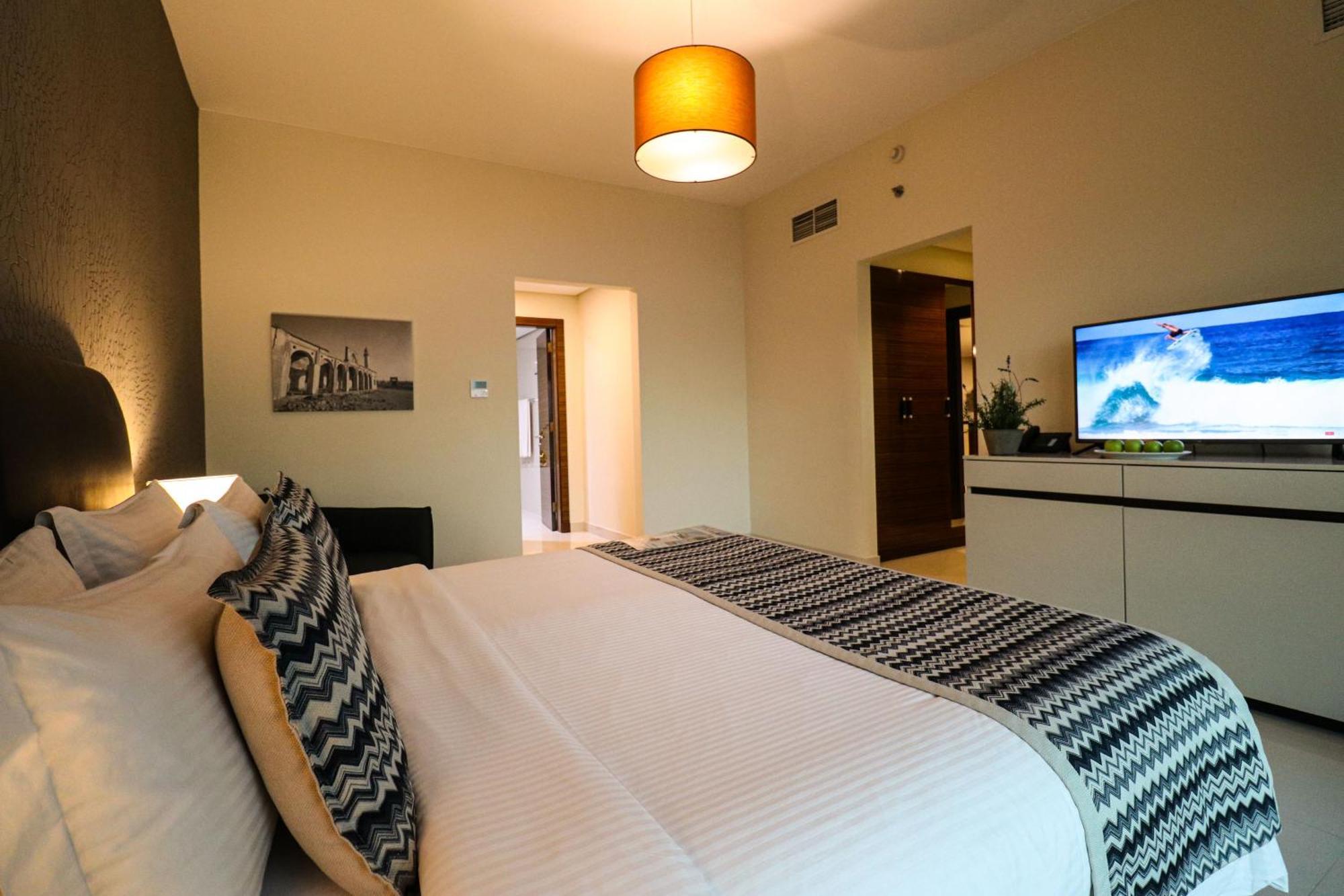 Lagoona Beach Luxury Resort And Spa Manama Eksteriør billede The photo shows a well-decorated hotel room. There is a large bed with white linens and decorative pillows featuring a zigzag pattern. On the wall beside the bed is a piece of artwork, and there is a modern television on a white furniture unit opposi
