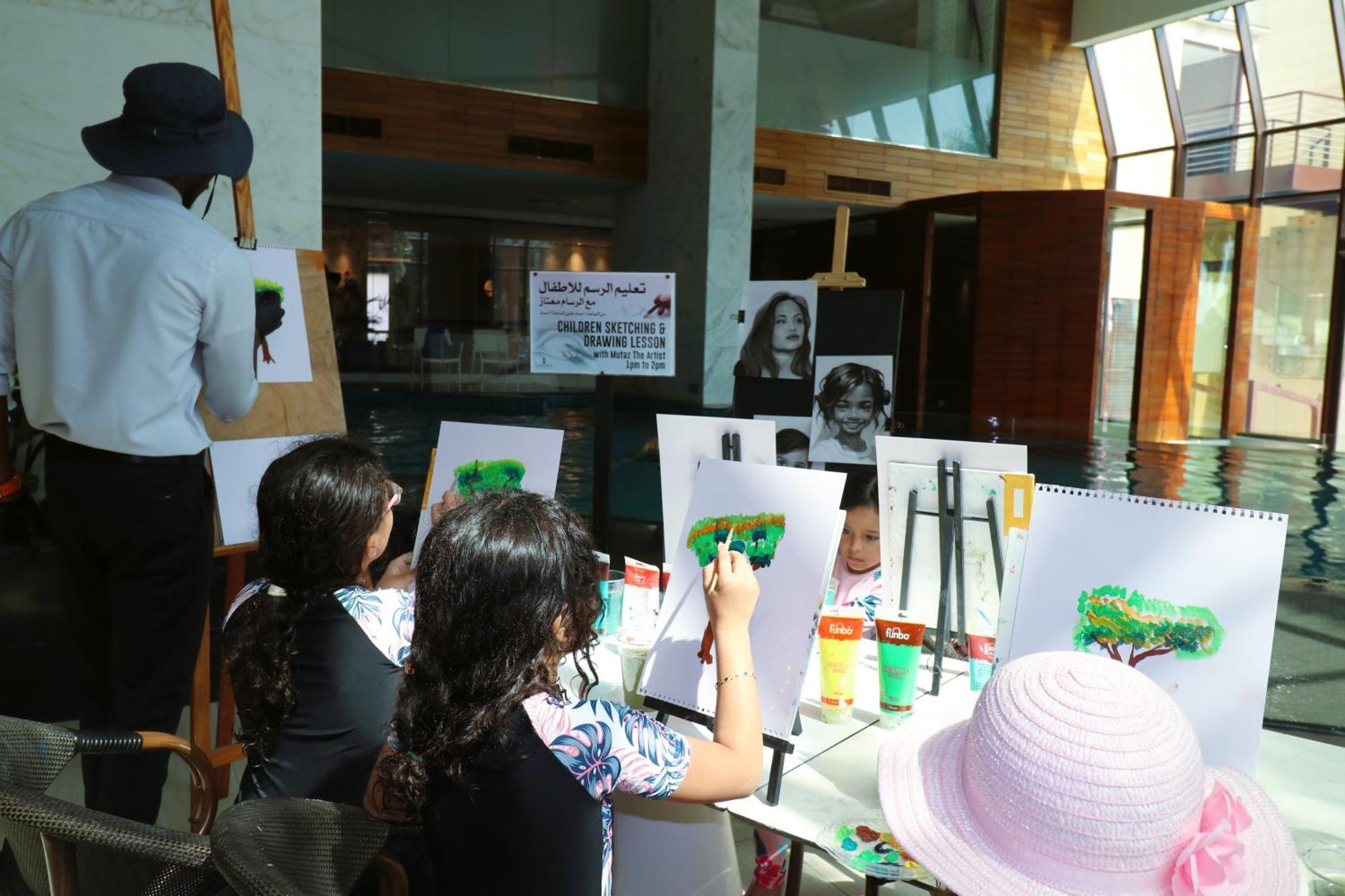 Lagoona Beach Luxury Resort And Spa Manama Eksteriør billede The photo shows a group of children and an adult engaged in an art activity near a pool area. The children are seated at a table, painting on canvases or paper, with a focus on trees, as indicated by the green paint they are using. An adult, wearing 