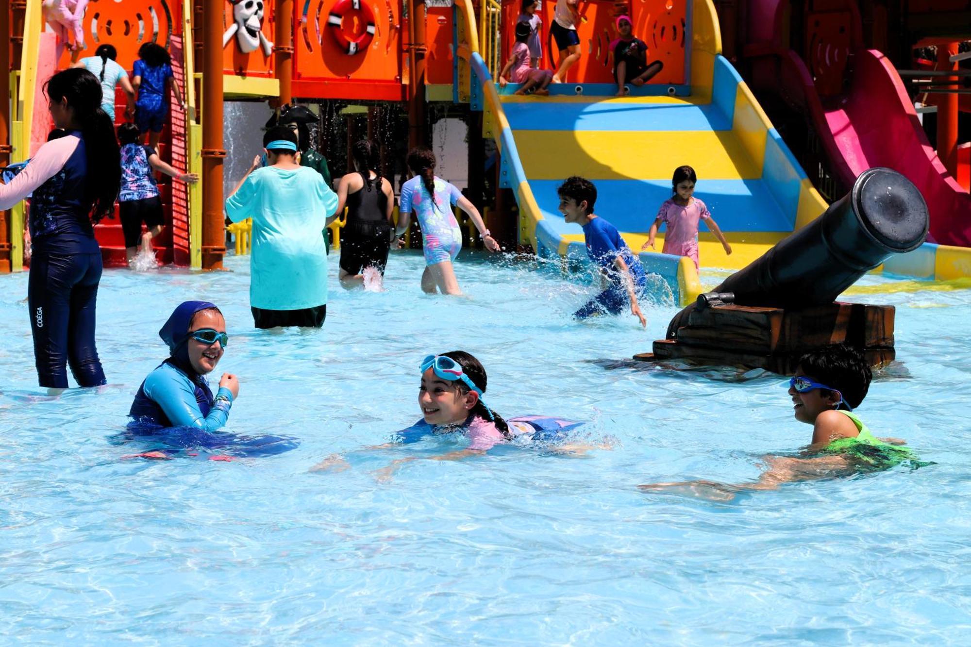 Lagoona Beach Luxury Resort And Spa Manama Eksteriør billede The photo shows a vibrant water park scene with several children playing in a shallow pool. Some are swimming and splashing around, while others are playing on colorful slides in the background. The atmosphere is lively and cheerful, with kids of var