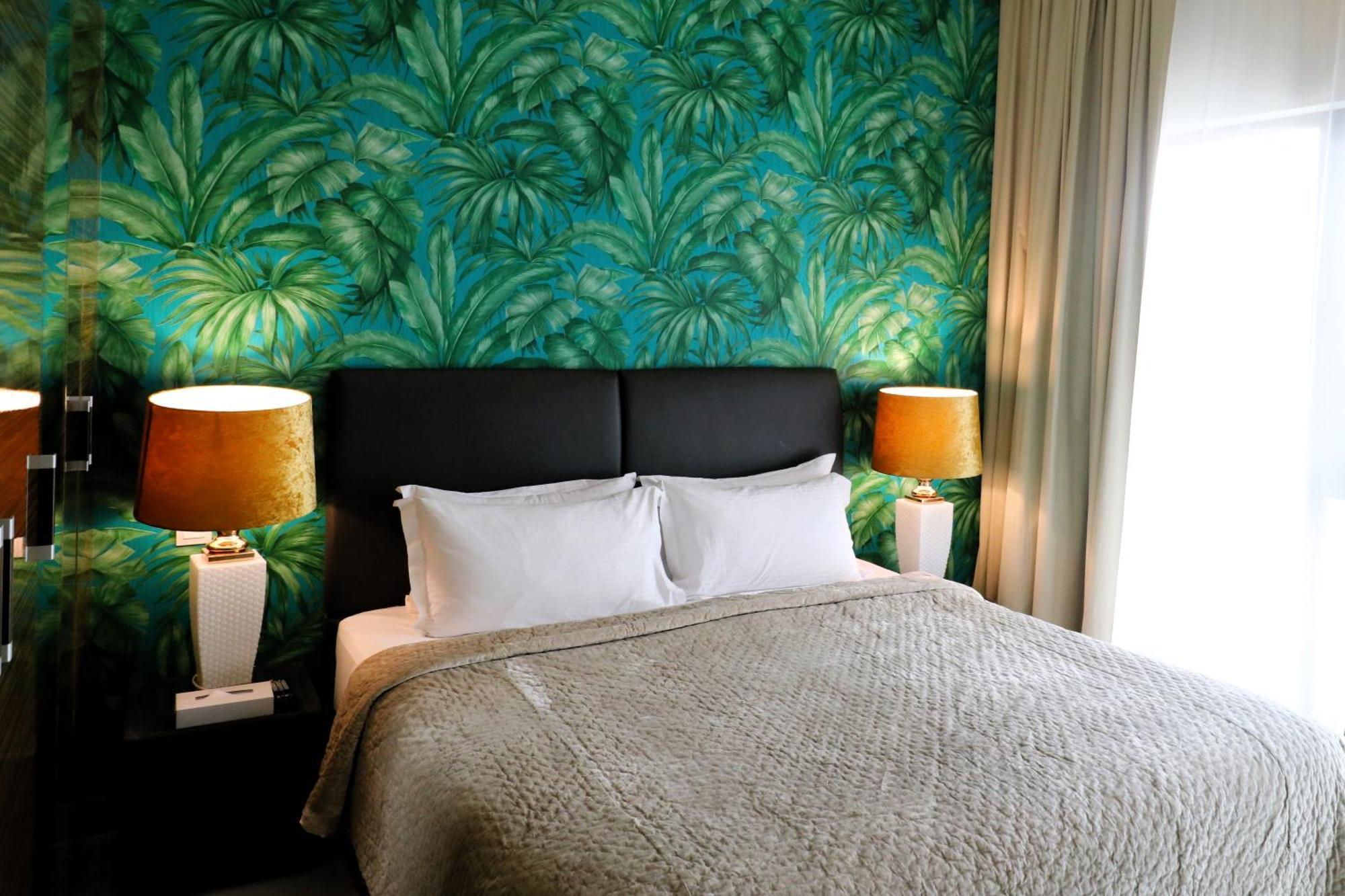 Lagoona Beach Luxury Resort And Spa Manama Eksteriør billede The photo shows a stylish bedroom featuring a large bed with white pillows and a gray blanket. The headboard is upholstered in black, providing a modern touch. Behind the bed, there is a vibrant green wallpaper with a tropical leaf pattern, adding a 