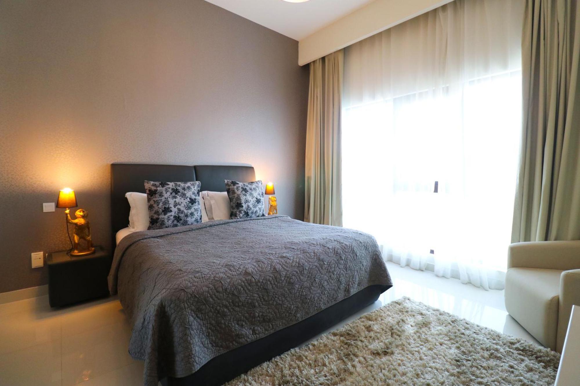 Lagoona Beach Luxury Resort And Spa Manama Eksteriør billede The photo shows a stylish bedroom with a modern design. It features a large bed with a dark gray bedspread and decorative pillows. On either side of the bed are two wooden nightstands, each with a lamp. There is a large window covered with light, flo